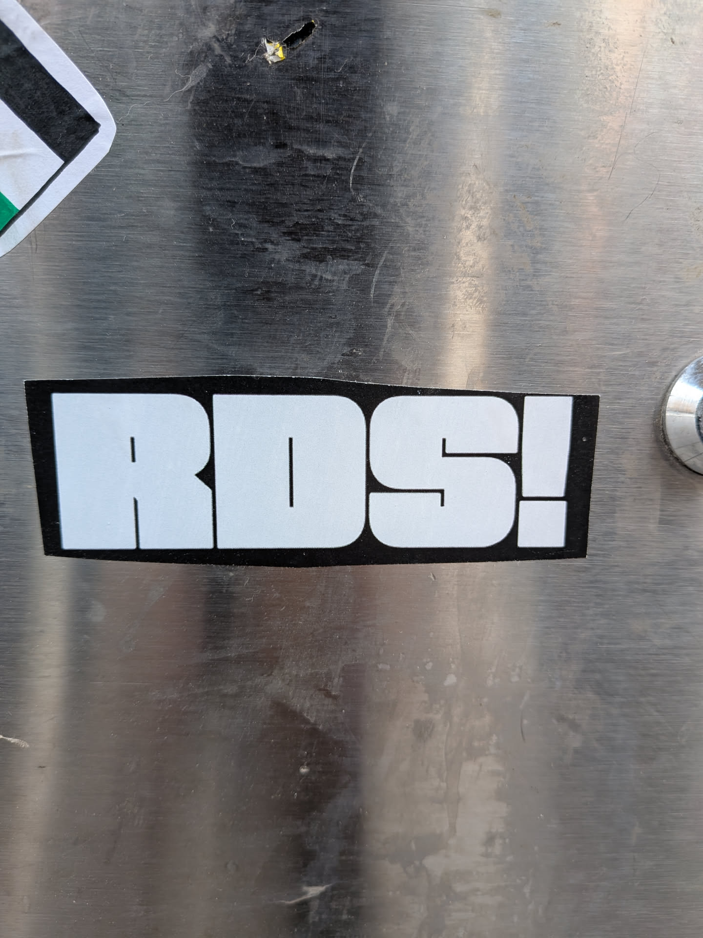 A rectangular sticker with a solid black background and slightly rounded corners. The sticker features the text "RDS!" in large, bold, white, sans-serif block letters. The contrast between the white text and the black background is stark, making the text highly prominent. The sticker is applied to a brushed metal surface, showing some wear and tear around its edges.