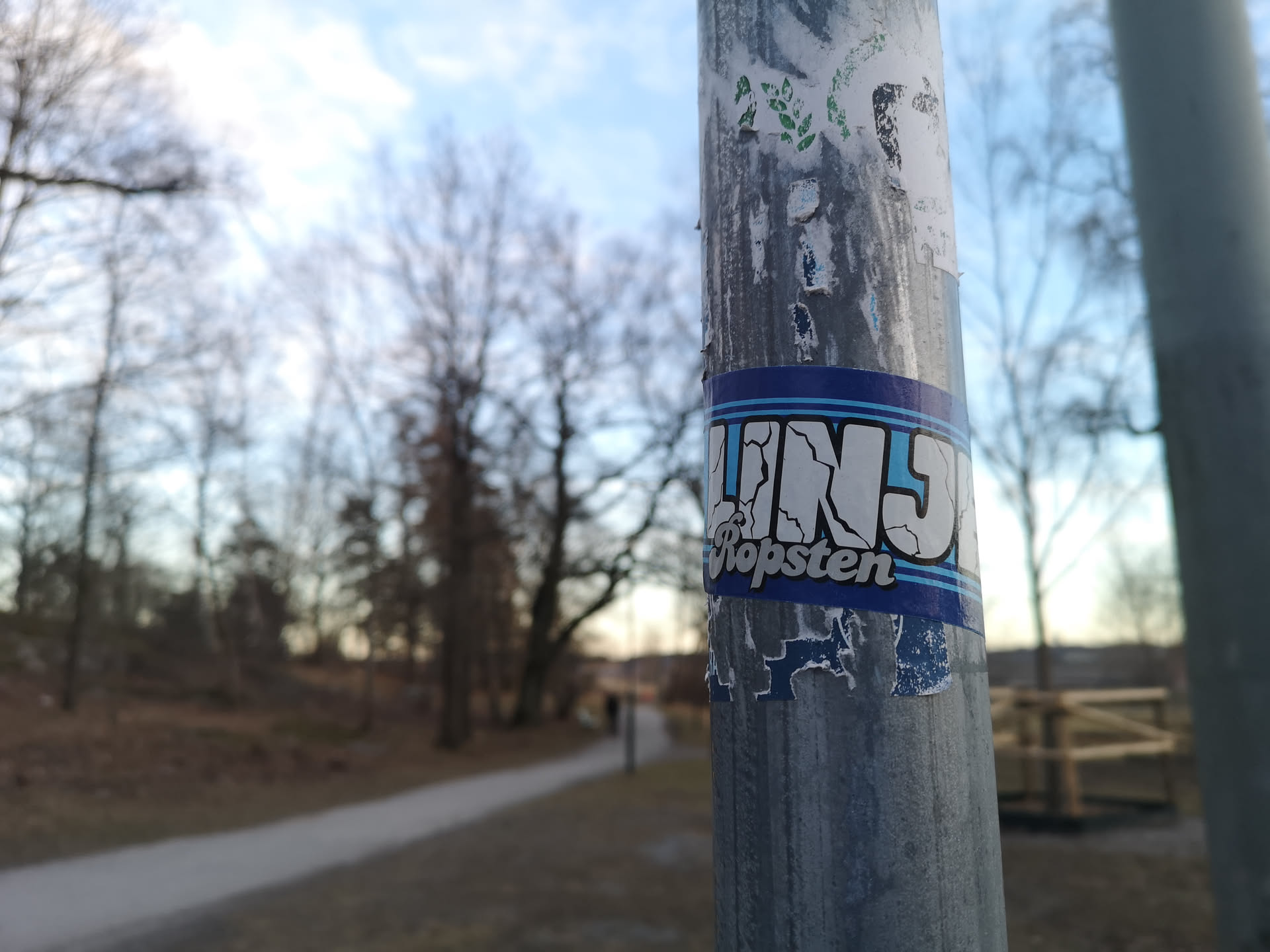 The sticker is rectangular with a dark blue background and a lighter blue border. The main text, 'TINS', is prominently displayed in large, white, blocky letters with a light blue outline or shadow. The letters have a distressed or cracked appearance, resembling a graffiti style. Below 'TINS', the word 'Ropsten' is written in a smaller, simpler, white sans-serif font, also with a light blue outline. The sticker shows signs of wear, with some peeling and fading, particularly along its bottom edge, and is affixed to a grey metal pole.