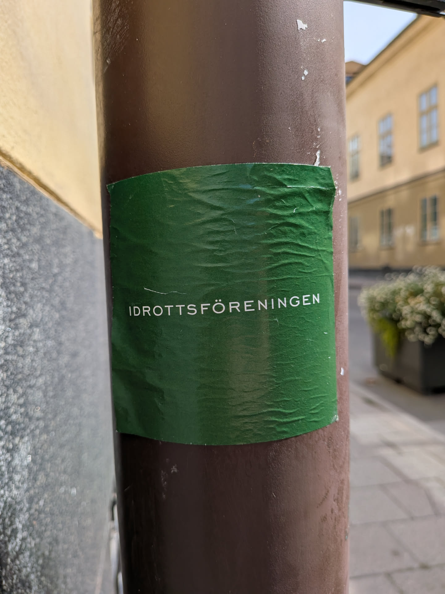 A wrinkled, rectangular green sticker affixed to a brown pole, featuring the white Swedish text 'IDROTTSFÖRENINGEN' in a clean sans-serif font.