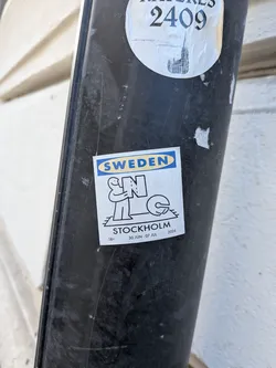 Square white sticker with an IKEA-style logo reading 'SWEDEN'. Features a black line art illustration of a figure holding the letter 'N' near a 'C' on a rug. Text at the bottom reads 'STOCKHOLM', '18+', and '30 JUN - 07 JUL 2026'.
