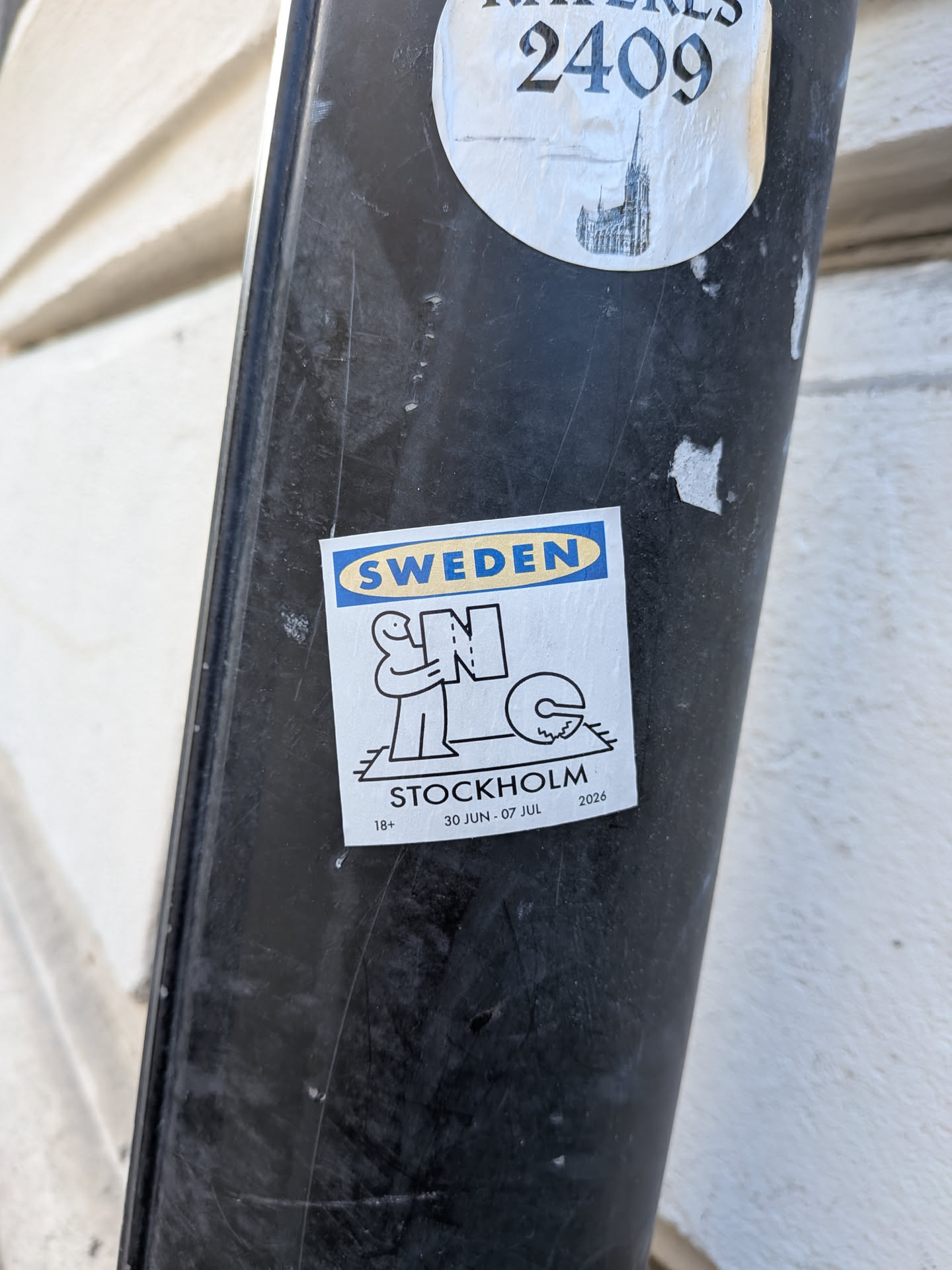 Square white sticker with an IKEA-style logo reading 'SWEDEN'. Features a black line art illustration of a figure holding the letter 'N' near a 'C' on a rug. Text at the bottom reads 'STOCKHOLM', '18+', and '30 JUN - 07 JUL 2026'.