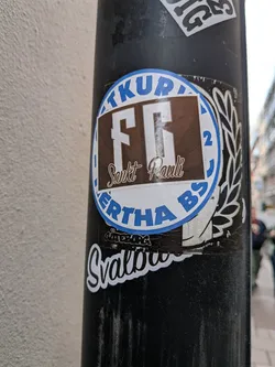 Street sticker A circular blue-and-white Hertha BSC sticker on a black pole, overlaid with a brown rectangular FC Sankt Pauli sticker. A smaller black 'Göteborg' sticker is visible underneath.