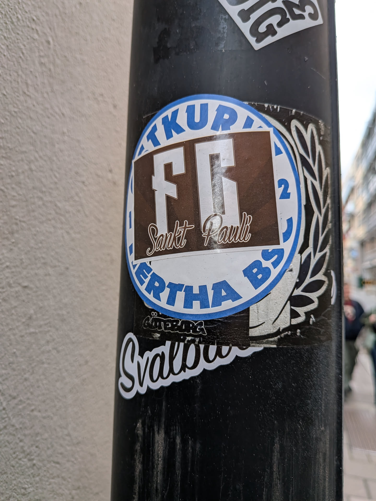 A circular blue-and-white Hertha BSC sticker on a black pole, overlaid with a brown rectangular FC Sankt Pauli sticker. A smaller black 'Göteborg' sticker is visible underneath.
