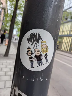 Street sticker A circular sticker featuring three stylized male figures on a white background. The figures are depicted in a cartoonish, somewhat gritty style. From left to right: The first figure has dark hair styled into a mohawk-like shape, a beard, and wears dark sunglasses, a black t-shirt, and grey pants, with visible arm tattoos. The central figure is bald with a prominent beard, an angry or stern expression, wearing a black t-shirt and grey pants, also with arm tattoos. The third figure has long blonde hair with a white headband, a beard, and wears a black t-shirt and grey pants. Above their heads are several abstract, black, dripping shapes, resembling stylized hair or a logo. The overall aesthetic suggests a band or a group with a heavy metal or rock influence.