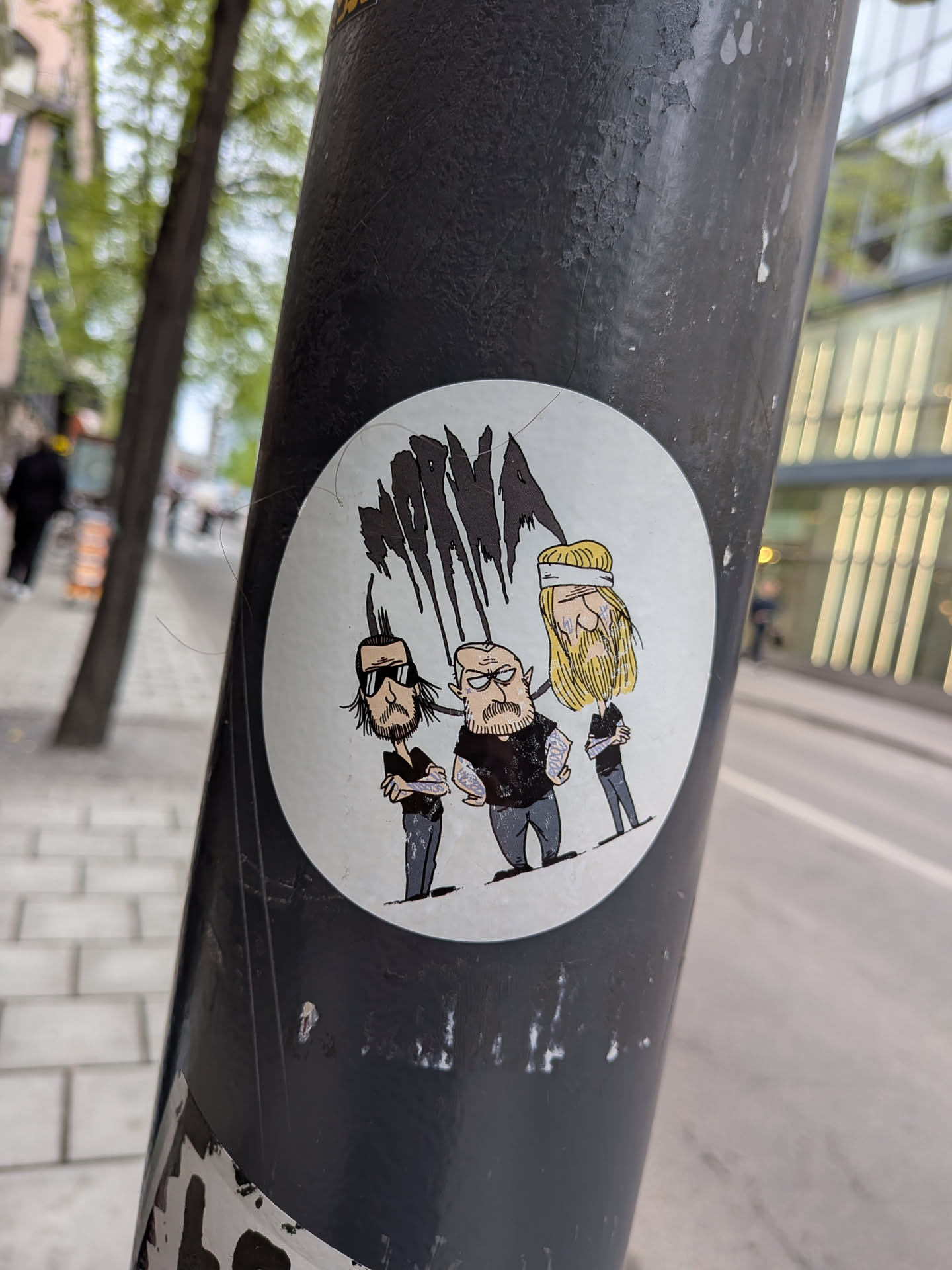 A circular sticker featuring three stylized male figures on a white background. The figures are depicted in a cartoonish, somewhat gritty style. From left to right: The first figure has dark hair styled into a mohawk-like shape, a beard, and wears dark sunglasses, a black t-shirt, and grey pants, with visible arm tattoos. The central figure is bald with a prominent beard, an angry or stern expression, wearing a black t-shirt and grey pants, also with arm tattoos. The third figure has long blonde hair with a white headband, a beard, and wears a black t-shirt and grey pants. Above their heads are several abstract, black, dripping shapes, resembling stylized hair or a logo. The overall aesthetic suggests a band or a group with a heavy metal or rock influence.