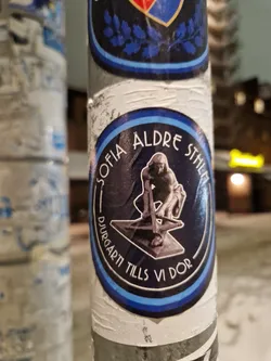 Street sticker A round sticker with a dark blue background and light blue border. It features an illustration of a figure sitting on a mechanism, possibly a statue. The text around the image translates to 'Sofia Aldre Stahl Djurgården until we die'.
