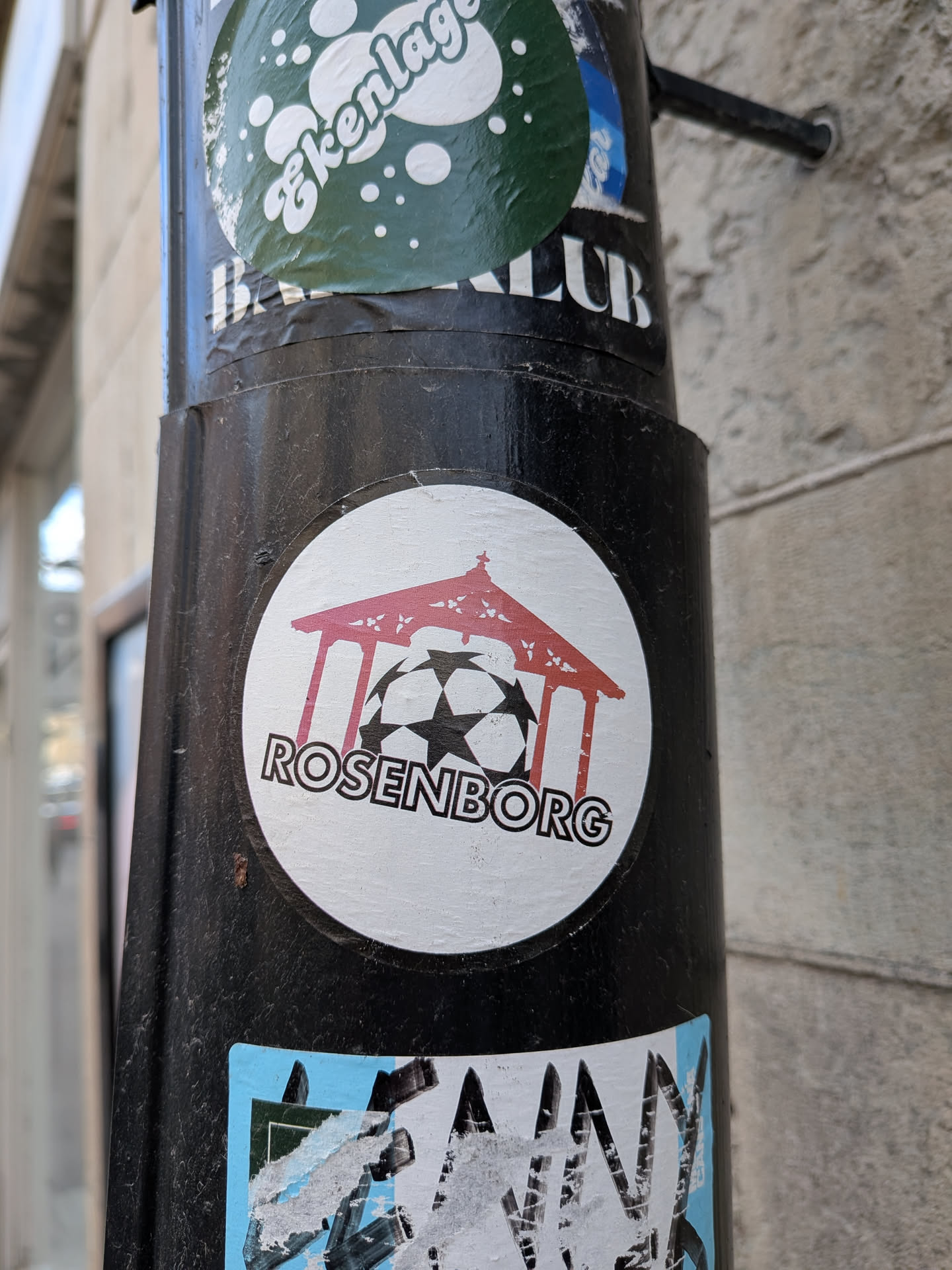 A circular white sticker featuring a stylized red pavilion or gazebo-like structure. Beneath this structure, a black and white soccer ball is depicted, resembling the UEFA Champions League ball with its characteristic star pattern. The word "ROSENBORG" is written in bold, black, sans-serif capital letters, curving along the bottom edge of the circle, partially overlapping the soccer ball.