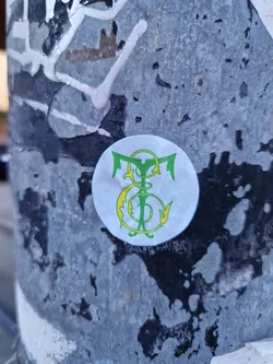 A round sticker with a monogram featuring intertwined letters or symbols in green and yellow on a white background.  The style is reminiscent of vintage or heraldic designs. It's affixed to a weathered surface.