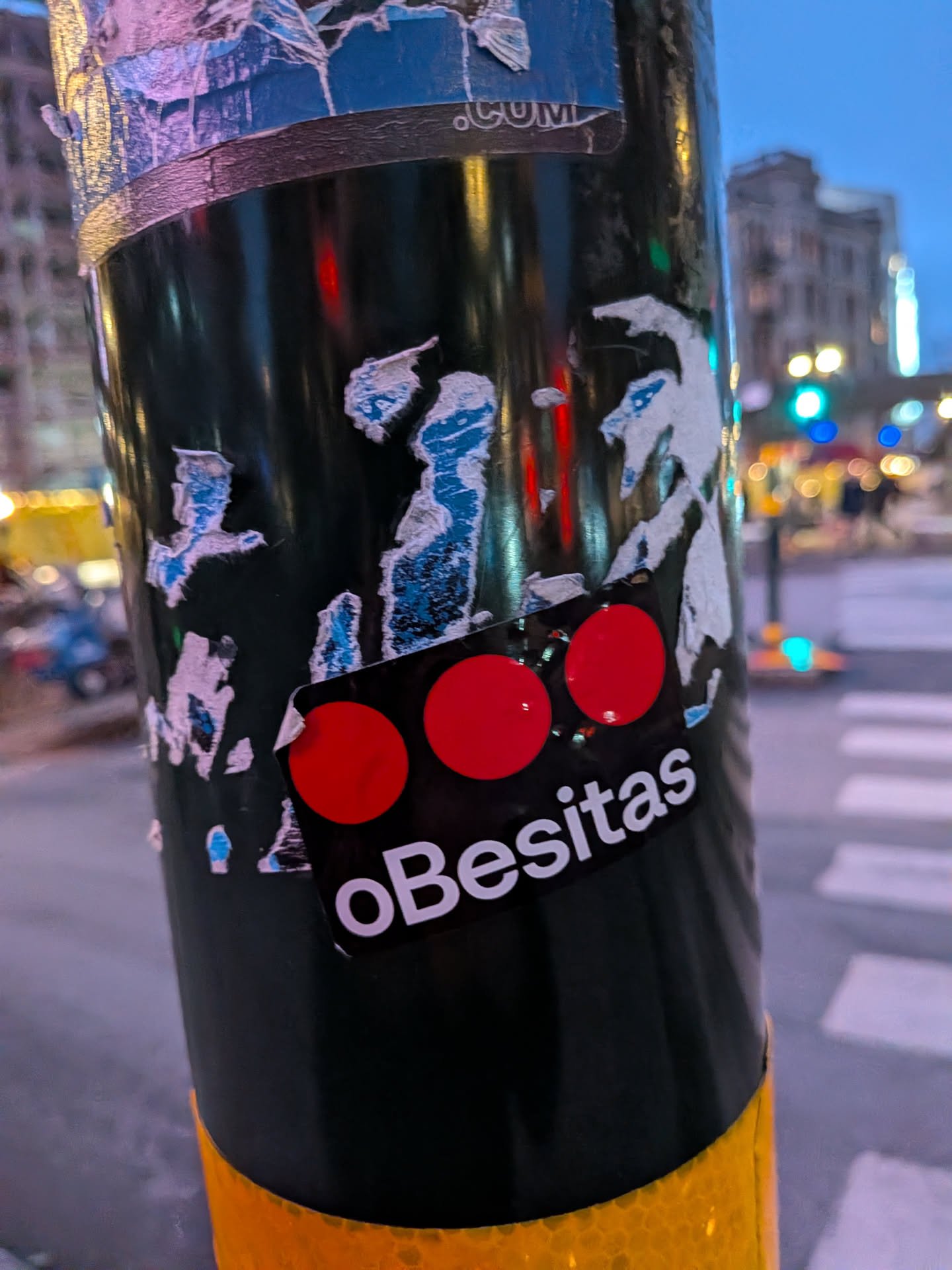 The sticker is rectangular with rounded corners and a black background. It features three prominent, solid red circles arranged horizontally in the upper portion. Below the circles, the word 'oBesitas' is written in a clean, white, sans-serif font. The sticker appears to be adhered to a dark, cylindrical object, with some peeling and wear visible around its edges, suggesting it has been exposed to the elements.