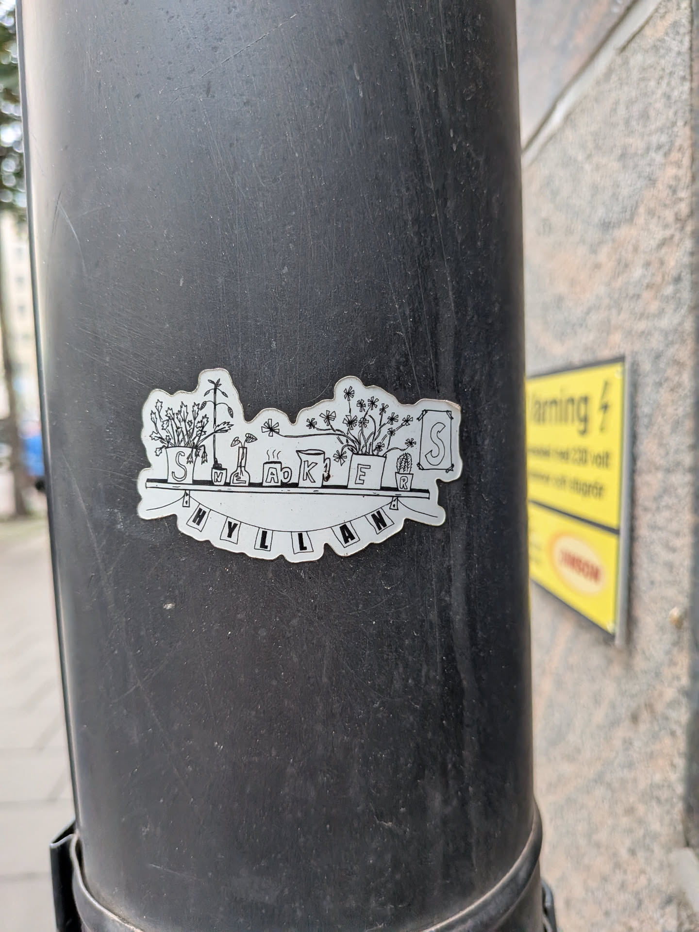 A die-cut, black and white sticker featuring a hand-drawn illustration of a shelf. On the shelf are various potted plants, a steaming mug, a pitcher, and a framed picture, each marked with a letter to spell 'SNEAKERS'. Hanging below the shelf is a banner that spells 'HYLLAN'.