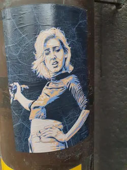 A sticker depicting a woman with short blonde hair, in a minimalist color scheme of beige and dark blue, set against a background resembling a city map.  The style is reminiscent of screen printing or stencil art.