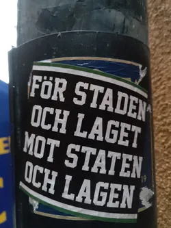 A black and white sticker with green and gold accents. The text is in Swedish and translates to "For the city and the team against the state and the teams". The number 19 is visible in the lower right corner. The sticker is affixed to a dark gray pole.