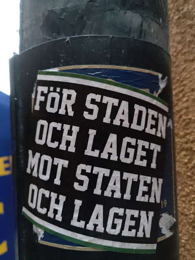 Street sticker A black and white sticker with green and gold accents. The text is in Swedish and translates to "For the city and the team against the state and the teams". The number 19 is visible in the lower right corner. The sticker is affixed to a dark gray pole.