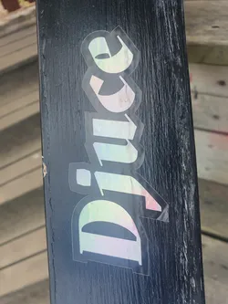 Street sticker This iridescent sticker features the word "Dive" in a stylized, holographic font.  The sticker has a slight rainbow effect, possibly due to a special printing technique or material. The sticker appears to be adhered to a dark-colored surface, perhaps a snowboard or skateboard. 