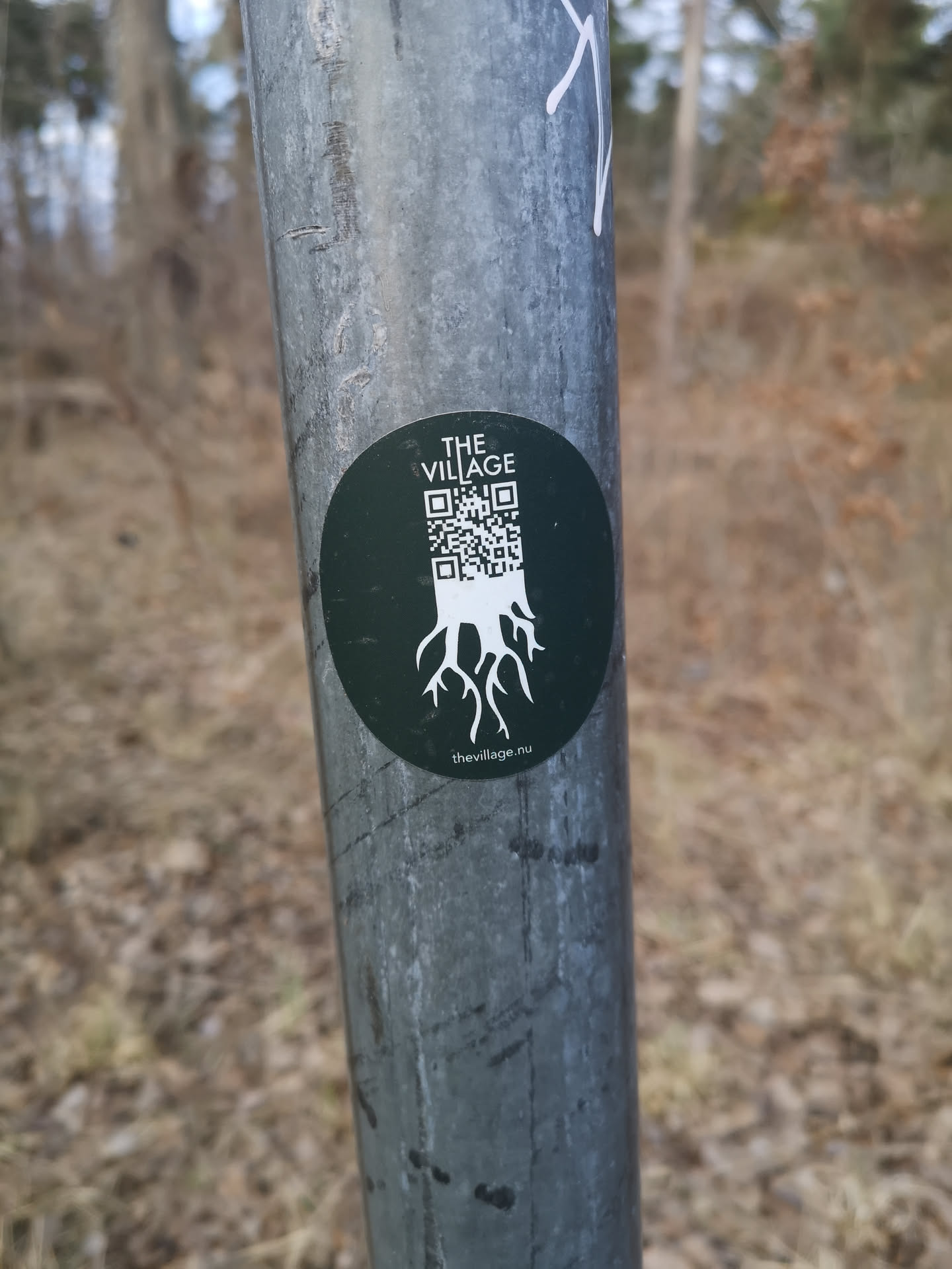 A circular sticker with a dark green background. The top features the white text 'THE VILLAGE' in a sans-serif font. Below this, a white QR code is integrated, with its lower portion transforming into a stylized depiction of white roots spreading downwards. At the very bottom, in smaller white text, is the website address 'thevillage.nu'.