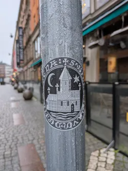 Street sticker The sticker is circular, featuring a monochromatic design primarily in black and white/grey. At its center is a detailed illustration of a multi-story building, strongly resembling an Ethiopian Orthodox Tewahedo Church, complete with a distinctive steeple topped with a cross. The building's architecture includes textured walls and arched openings. Below the church, wavy lines suggest water or a river. Within the circular frame, above the church, a crescent moon is positioned on the left and a five-pointed star on the right. Additional small crosses, characteristic of Coptic or Ethiopian design, are visible near the church. The entire central image is encircled by a thick border containing text rendered in white against a black background. The script appears to be Ge'ez, an ancient Ethiopic script. The sticker is affixed to a wet, metallic pole, with water droplets visible on its surface, indicating it is a physical object in the environment.