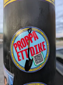 Street sticker The central sticker is circular with a light blue background. It features a stylized black shovel with a brown handle pointing diagonally downwards. Bold, red text across the sticker reads 'PROPPA ETT DIKE'. Around the circumference of the sticker, in yellow text on a blue band, are the words 'ÅTERSTÄLL VÅTMARKER' repeated. A green-bordered square containing a QR code is positioned in the lower right quadrant of the sticker.