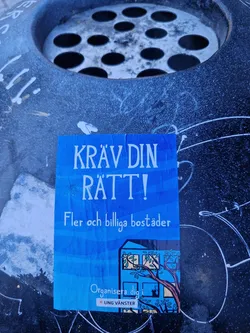 Street sticker A blue sticker with the text "Kråv din rätt!" (Demand your right!) in large letters. Below it, in smaller text, it says "More and cheaper housing." There is an illustration of a house with trees and a cat in the window. At the bottom, it says "Organize yourself in Ung Vänster" (Young Left).