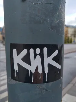 Black and white sticker with the word Khik written in a graffiti style with dripping paint effect. The sticker is placed on a grey pole.