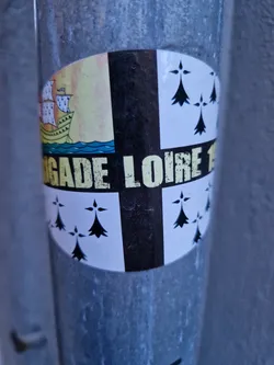 Round sticker with a design that looks like a heraldic emblem. It's divided into quadrants, with a ship on one side, ermine spots on two others, and a central black section with partially visible text. The visible text reads 'GADE LOIRE 1'.