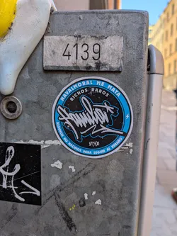 Street sticker The sticker is circular, featuring a striking design primarily in black, bright blue, and white. It has a prominent blue outer ring. The center of the sticker contains a highly stylized, graffiti-style word in white with a black outline, which appears to be 'Familia' or a similar word, accompanied by a small white lightning bolt symbol to its right. Along the top inner edge of the blue ring, the white text reads 'NAVALMORAL ME MATA' and 'BICHOS RAROS'. The bottom inner edge of the blue ring displays the white phrase 'ALGUNOS NO NACIMOS PARA SEGUIR AL REBAÑO'. A small numerical sequence '10500' is visible in white at the very bottom center, beneath the main stylized text. The sticker is applied to a textured, grey metal surface, showing some signs of wear.