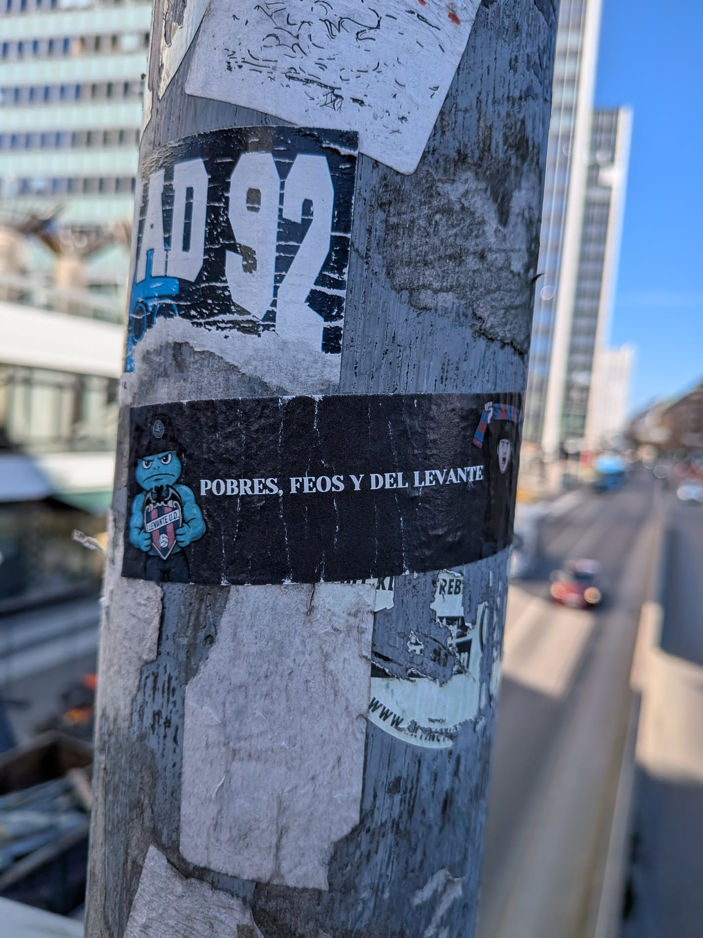 The sticker is primarily black with white text and a blue character. The top section features large, distressed white letters 'AD 92' on a black background. Below this, the main part of the sticker is a black rectangle. On the left side, a light blue, cartoonish character with a grumpy expression is depicted, wearing a black hat and a red and blue striped scarf. The character holds a shield or badge that clearly reads 'LEVANTE U.D.' in white text. To the right of the character, the phrase 'POBRES, FEOS Y DEL LEVANTE' is written in white capital letters. The sticker shows signs of wear and tear, with some peeling and texture visible on the edges and surface.