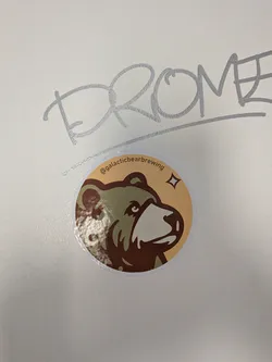 A circular sticker featuring a stylized illustration of a bear's head, facing right. The bear has dark brown fur with a lighter tan snout and a dark eye. The background of the sticker is a warm, light tan color, with a small, four-pointed star or sparkle icon in the upper right. Along the top curve of the sticker, the text "@galacticbearbrewing" is written in a dark brown font.