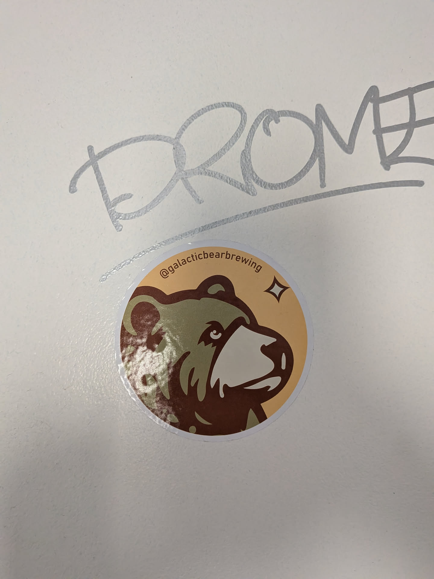 A circular sticker featuring a stylized illustration of a bear's head, facing right. The bear has dark brown fur with a lighter tan snout and a dark eye. The background of the sticker is a warm, light tan color, with a small, four-pointed star or sparkle icon in the upper right. Along the top curve of the sticker, the text "@galacticbearbrewing" is written in a dark brown font.
