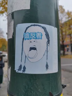 Street sticker This sticker shows a close-up of a person's face, depicted in a minimalist style with dark hair styled in braids.  The eyes are covered by a rectangular shape with Chinese characters. The mouth is open in a shout or yell. The overall aesthetic is bold and slightly edgy.