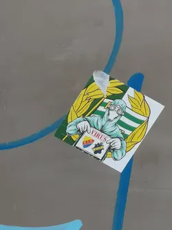 Street sticker A square-shaped sticker featuring a cartoonish figure clad in a light blue/teal hazmat suit, complete with goggles and gloves, against a background of horizontal green and white stripes. The figure is holding a white sign that reads 'VIRUS' in prominent red letters at the top. Below the text are two distinct football club crests: one with 'D.I.F.' in yellow on a red background, and another with 'A.I.K.' in yellow on a dark blue/black background. The central image is framed by two large, stylized yellow leaf or wheat stalk designs. The sticker shows minor wear, particularly a slight peel at the top left corner.