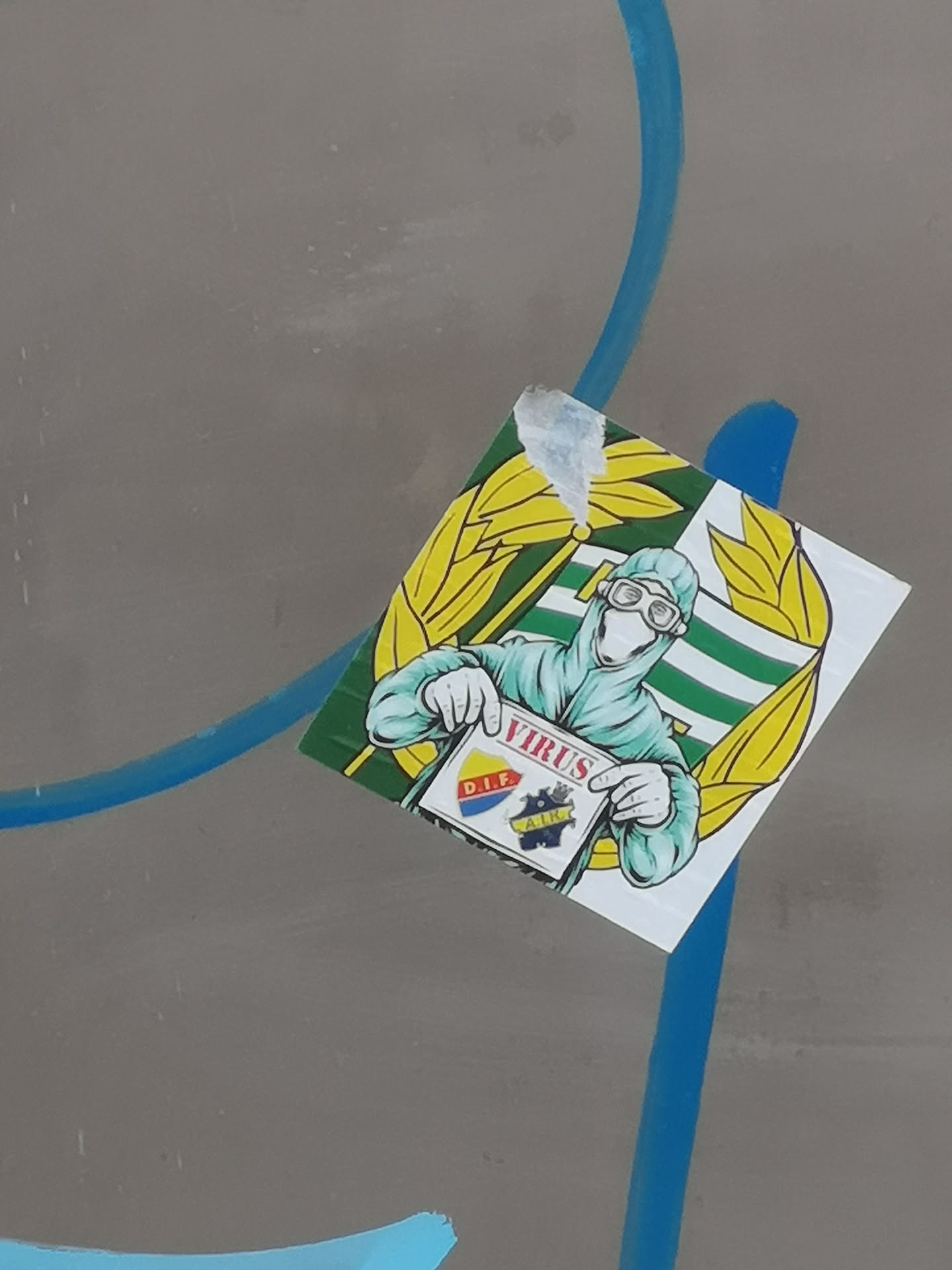 A square-shaped sticker featuring a cartoonish figure clad in a light blue/teal hazmat suit, complete with goggles and gloves, against a background of horizontal green and white stripes. The figure is holding a white sign that reads 'VIRUS' in prominent red letters at the top. Below the text are two distinct football club crests: one with 'D.I.F.' in yellow on a red background, and another with 'A.I.K.' in yellow on a dark blue/black background. The central image is framed by two large, stylized yellow leaf or wheat stalk designs. The sticker shows minor wear, particularly a slight peel at the top left corner.