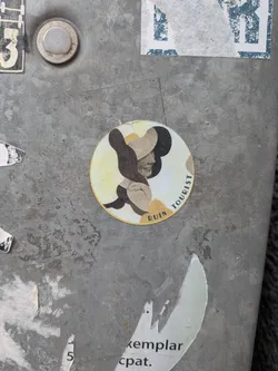Street sticker A circular sticker featuring a stylized profile of a woman facing right, wearing a large, wide-brimmed hat. The image is rendered in a collage or cut-out style, using abstract shapes in muted earthy tones of dark brown, black, beige, and pale yellow. Her face is light-colored with a subtle smile, and her hat is composed of dark brown and beige sections. The background within the circle is a pale yellow. The words 'RUIN' and 'TOURIST' are curved along the bottom-left and right edges of the circle, respectively.
