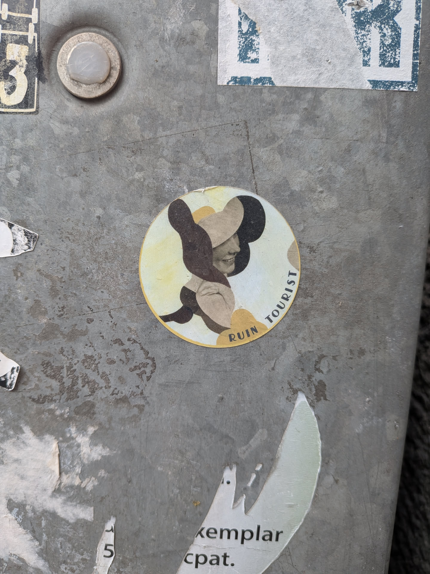 A circular sticker featuring a stylized profile of a woman facing right, wearing a large, wide-brimmed hat. The image is rendered in a collage or cut-out style, using abstract shapes in muted earthy tones of dark brown, black, beige, and pale yellow. Her face is light-colored with a subtle smile, and her hat is composed of dark brown and beige sections. The background within the circle is a pale yellow. The words 'RUIN' and 'TOURIST' are curved along the bottom-left and right edges of the circle, respectively.