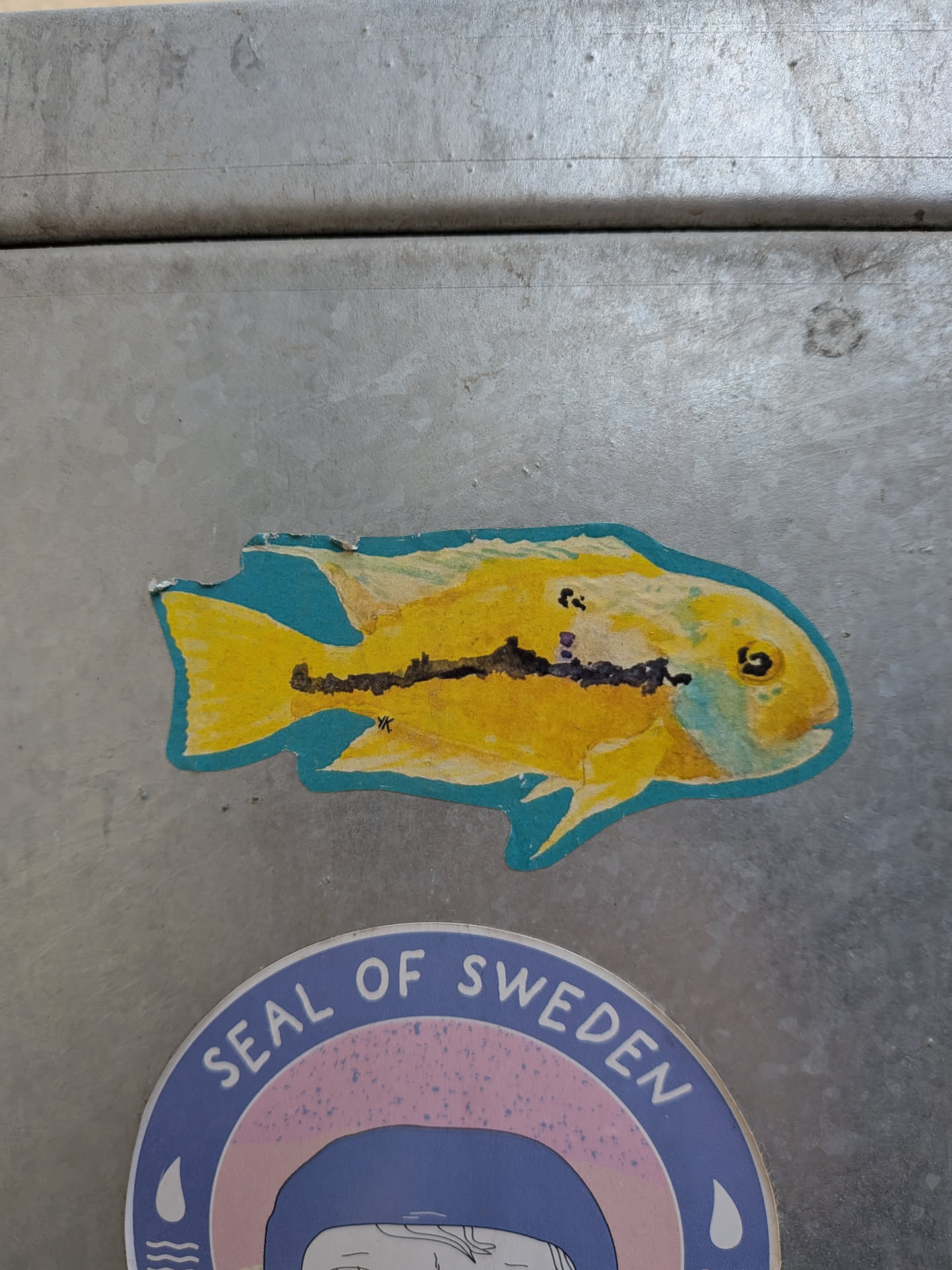 A die-cut sticker featuring a bright yellow fish, likely a cichlid, with a distinct blue outline. The fish's body is predominantly a vivid yellow, accented by a prominent horizontal black stripe or pattern running along its midsection, and a few smaller black spots on its dorsal area. Its eye is black with a yellow iris, and there are subtle hints of light blue or greenish coloration around the gills and head. Near the tail, the initials 'YK' are visible in black. The sticker has a textured appearance, suggesting it might be a print of a watercolor or colored pencil drawing. The edges show minor wear and slight peeling, particularly at the top.