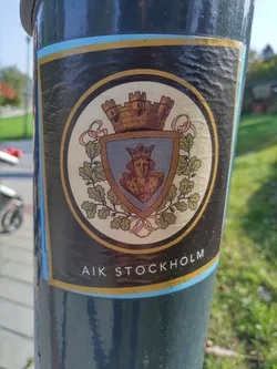 Street sticker A sticker featuring the emblem of AIK Stockholm. The emblem depicts a crowned woman's head within a shield, surrounded by oak leaves and a castle on top. The text 'AIK STOCKHOLM' is visible at the bottom.