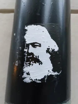 A black and white sticker featuring a stylized portrait of Karl Marx. The sticker is affixed to a dark-colored cylindrical surface, possibly a pole or pipe.