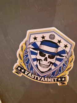 Street sticker This sticker features a skull wearing a hat, surrounded by a shield with stars.  The text at the bottom reads "VASTYARNET". The style is graphic and bold, with a combination of blue, white, and yellow colors.