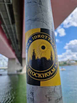 Street sticker A yellow and black circular sticker featuring a stylized cityscape of Stockholm. The text around the circle reads "Anna Idrottsklubben Stockholm". The design is simple yet striking, with a sunburst graphic behind the city skyline. The sticker is affixed to a metal pole.