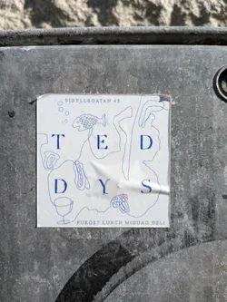 Street sticker A rectangular white sticker with blue line art and text, affixed to a dark, textured surface. The sticker features the name 'T E D D Y S' in large, spaced-out blue capital letters, arranged somewhat organically amidst abstract blue line drawings. These drawings include a stylized fish, a wine glass, small circles (possibly bubbles), and various organic, blob-like shapes. At the top, 'SIBYLLEGATAN 43' is written in smaller blue text, and at the bottom, 'FUKOST LUNCH MIDDAG DELI' is also in smaller blue text. The sticker shows signs of wear, with visible creases and slightly curled edges, indicating it's a physical sticker that has been exposed to elements.