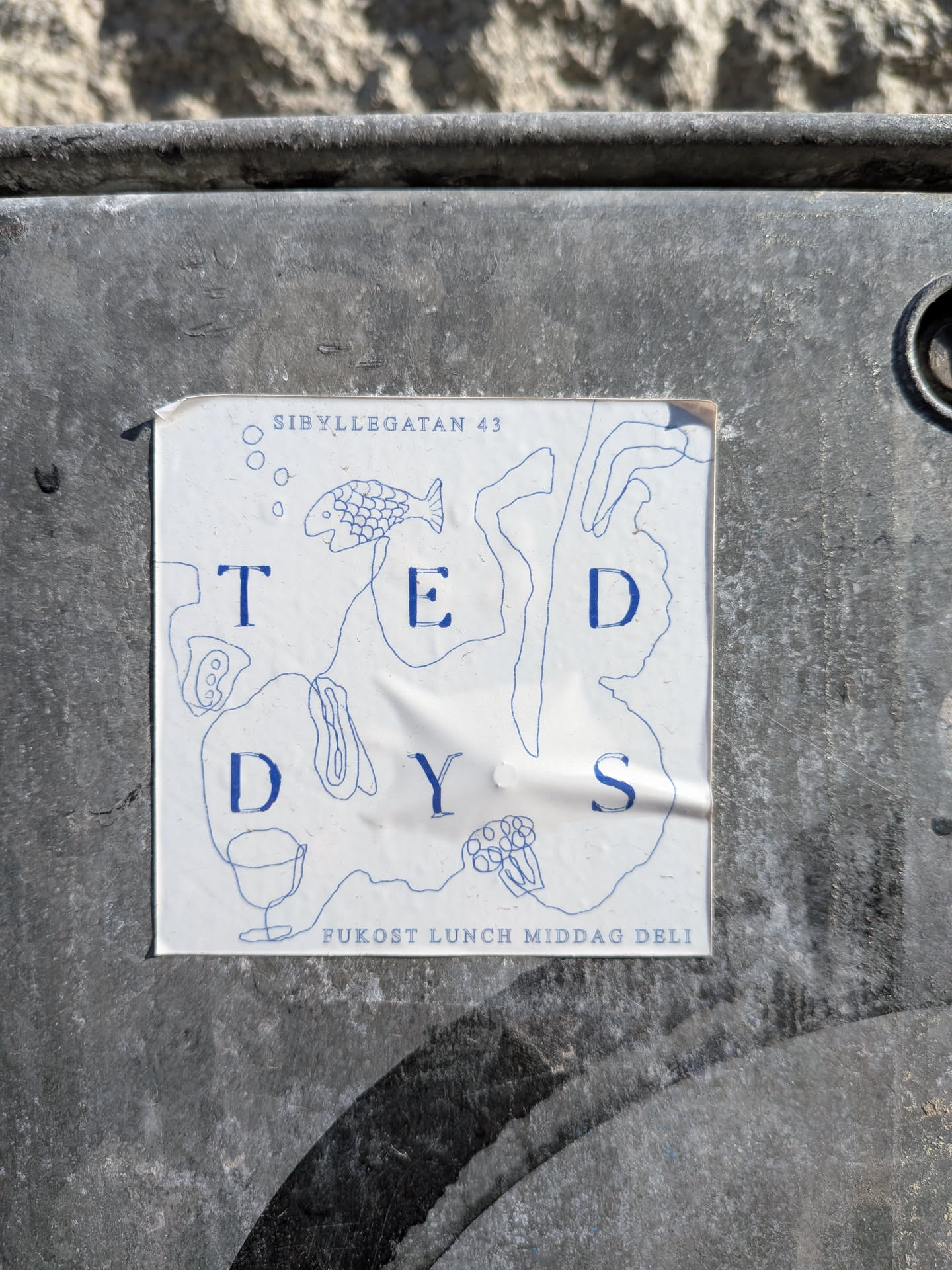 A rectangular white sticker with blue line art and text, affixed to a dark, textured surface. The sticker features the name 'T E D D Y S' in large, spaced-out blue capital letters, arranged somewhat organically amidst abstract blue line drawings. These drawings include a stylized fish, a wine glass, small circles (possibly bubbles), and various organic, blob-like shapes. At the top, 'SIBYLLEGATAN 43' is written in smaller blue text, and at the bottom, 'FUKOST LUNCH MIDDAG DELI' is also in smaller blue text. The sticker shows signs of wear, with visible creases and slightly curled edges, indicating it's a physical sticker that has been exposed to elements.