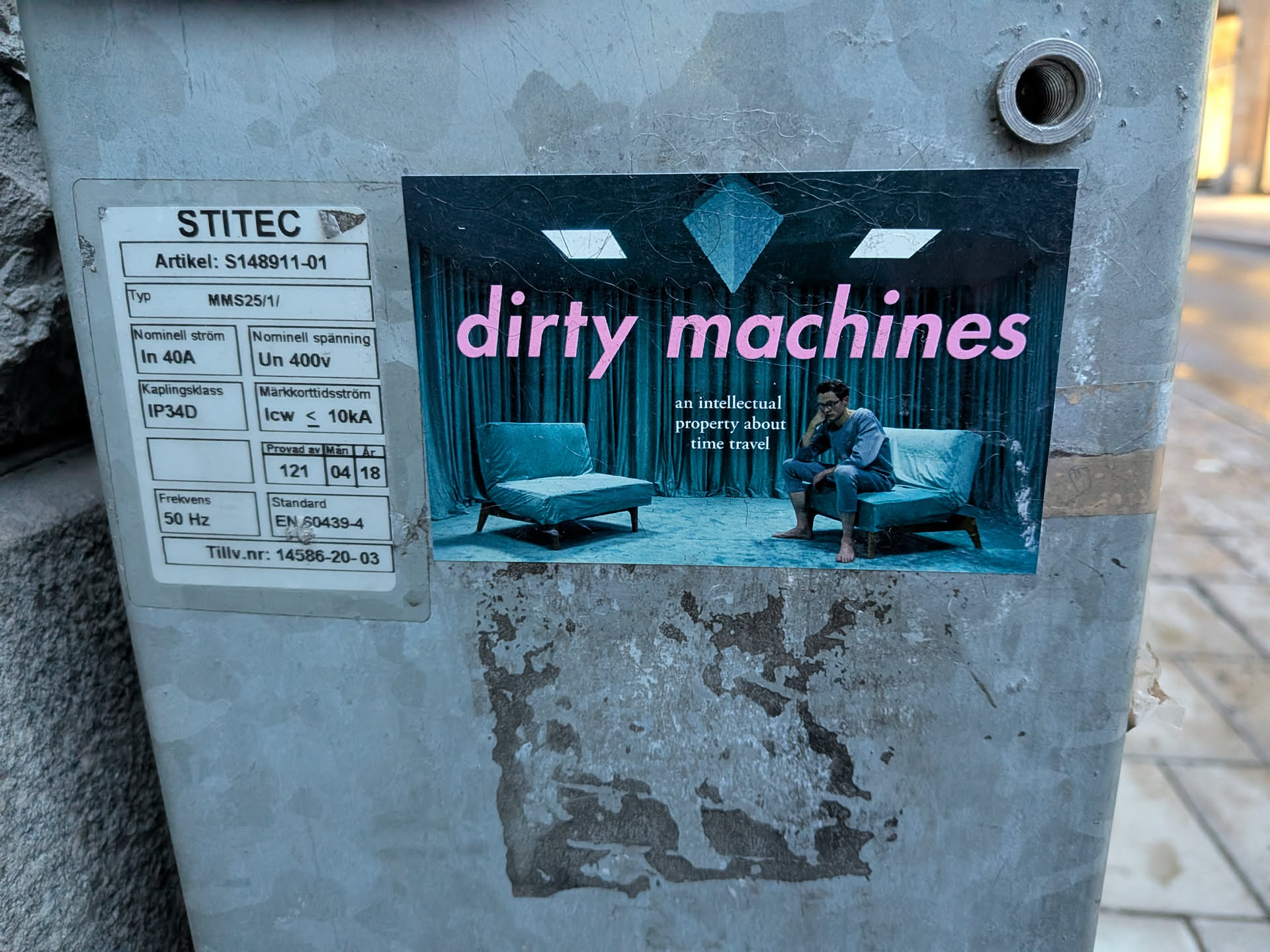 The sticker features a surreal scene with a dark, teal-colored backdrop of draped curtains and a diamond-shaped geometric element at the top. In the foreground, two minimalist, teal-colored chairs are placed on a dark, textured floor. A person in a grey outfit is seated on one of the chairs, looking contemplative. The text 'dirty machines' is prominently displayed in a large, pink, sans-serif font across the upper portion of the sticker, with smaller text below reading 'an intellectual property about time travel'.
