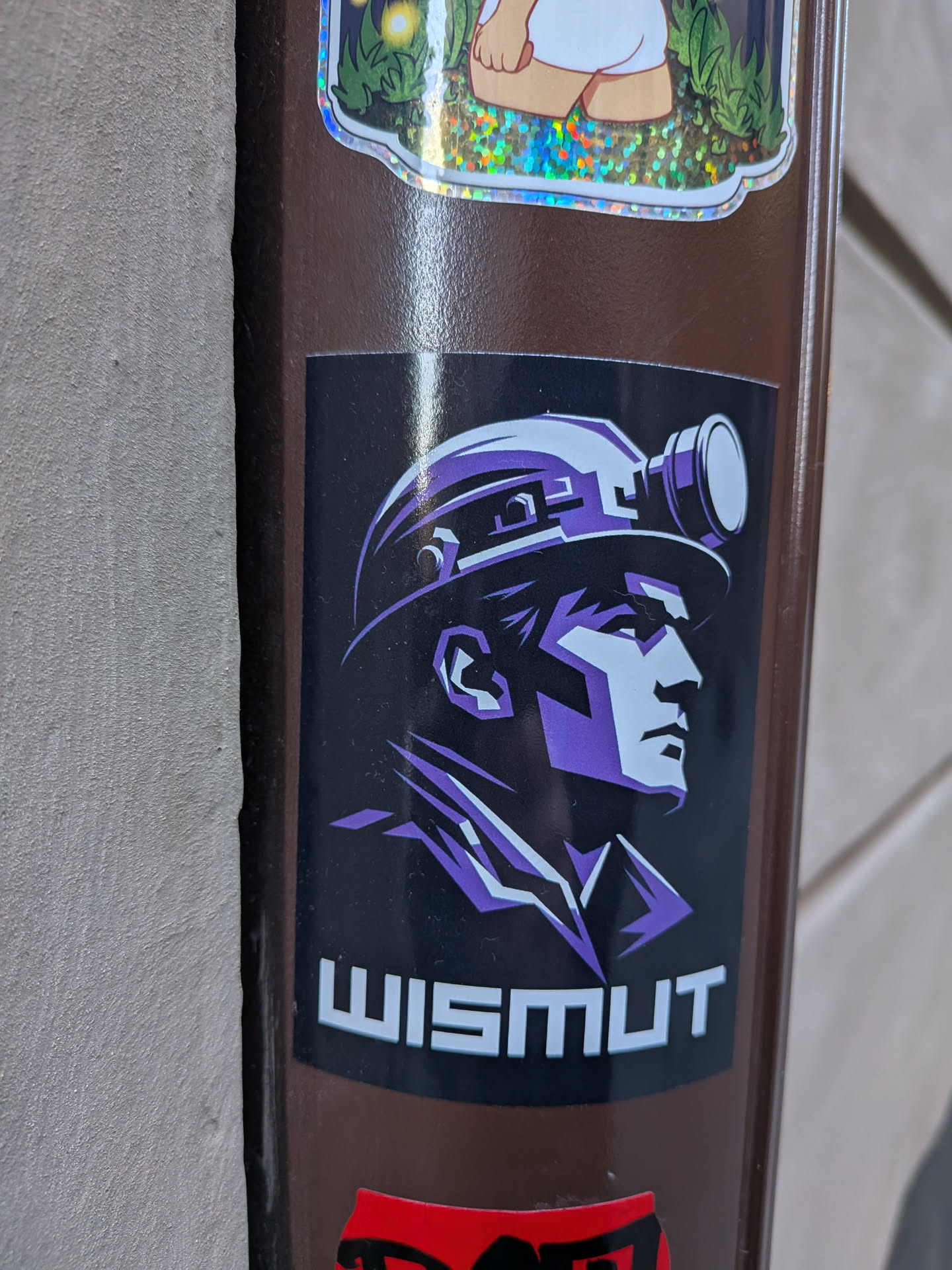 Street sticker The sticker features a stylized profile portrait of a person, likely a miner, facing towards the right. They are depicted wearing a hard hat with a prominent, circular headlamp on the front. The artwork uses a limited color palette, primarily shades of purple and white/light grey against a solid black background, creating a strong, graphic, and almost monochromatic effect. The lines are sharp and angular, giving the figure a determined and rugged appearance. Below the portrait, the word "WISMUT" is written in bold, white, sans-serif capital letters.