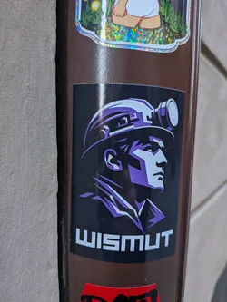 Street sticker The sticker features a stylized profile portrait of a person, likely a miner, facing towards the right. They are depicted wearing a hard hat with a prominent, circular headlamp on the front. The artwork uses a limited color palette, primarily shades of purple and white/light grey against a solid black background, creating a strong, graphic, and almost monochromatic effect. The lines are sharp and angular, giving the figure a determined and rugged appearance. Below the portrait, the word "WISMUT" is written in bold, white, sans-serif capital letters.