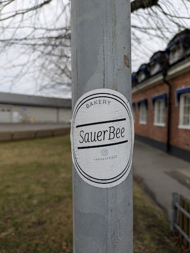 Street sticker A round sticker adhered to a gray pole, advertising "SauerBee Bakery". The sticker is white with a black border and black text. The text is arranged in a circular pattern, with "BAKERY" at the top, "SauerBee" in the middle, and the Instagram handle "@THESAUERBEE" at the bottom.