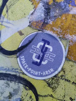 Street sticker A round sticker with a logo featuring overlapping letters 'S', 'P', and 'A', possibly an abbreviation for Space Point ArsN. The sticker is light purple with white text. It appears to be affixed to a wall with other graffiti.