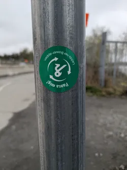 A round green sticker is affixed to a weathered metal pole. The sticker has the text "Ladda ner Bower-appen." and "Panta mig!" in a circular arrangement around an icon resembling a stylized recycling symbol. The sticker is slightly faded and shows signs of outdoor exposure. The pole is grey with vertical striations and minor surface imperfections. The background is blurred.