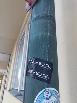 A black and white sticker with the words "NEW BLACK" repeated twice, along with a website address. The sticker appears to be placed on a dark-green metal pole.