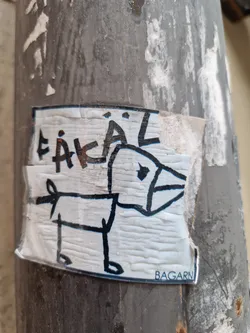 This sticker features a simple drawing of a dog, reminiscent of a child's artwork. The word "ÄKAL" is written above the dog in a stylized manner.  The sticker is affixed to a weathered surface, and shows signs of wear and tear. The word "BAGARN" is visible near the bottom.