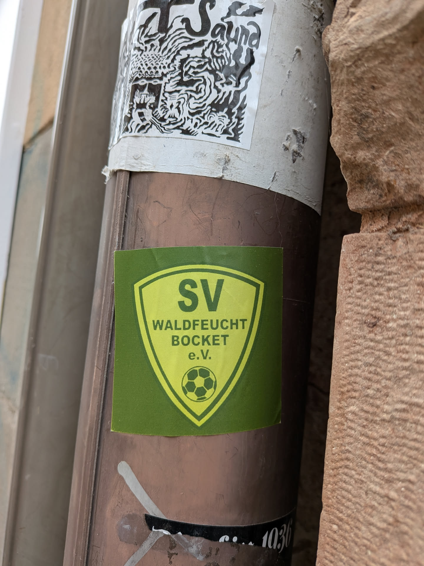 The sticker is a rectangular shape with a slightly textured, matte finish, primarily in a dark green color. Centered on it is a shield-shaped emblem in a lighter, lime-green or yellowish hue, outlined in a darker green. Inside the shield, the letters "SV" are prominently displayed at the top in dark green. Below "SV", the text "WALDFEUCHT BOCKET" is written in two lines, also in dark green. At the bottom of the shield, "e.V." is written, and beneath that, a stylized football (soccer ball) icon is depicted. The overall design is that of a traditional sports club logo.