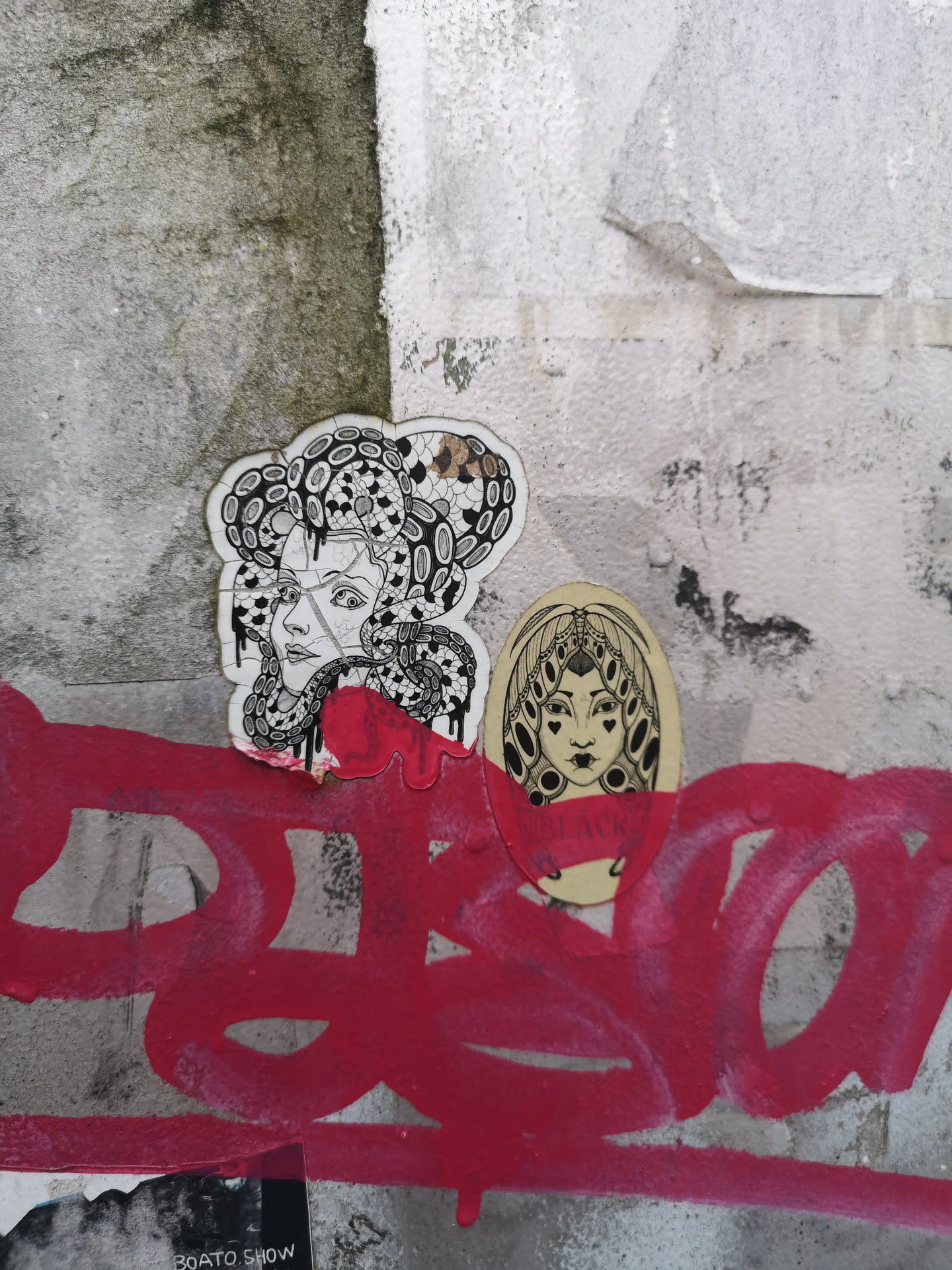 The image features two distinct, hand-drawn style stickers applied to a textured, grey and white wall, partially covered by vibrant red graffiti. The left sticker, larger and irregularly shaped, depicts a woman's head and shoulders in profile, looking to the left. Her hair is intricately detailed with snakes and swirling patterns, reminiscent of Medusa, rendered in black and white line art with subtle shading. Red paint drips from the bottom edge of this sticker, blending with the graffiti below. The right sticker, smaller and oval-shaped, presents a woman's face looking directly forward. She has stylized eyes with heart-shaped pupils and an elaborate, symmetrical headdress or hairstyle. This sticker is also executed in black and white line art. A band of red paint from the graffiti covers the lower part of her face, partially obscuring some text.